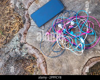 Un téléphone et un nœud de quatre paires d'écouteurs intra-auriculaires filaires rose, bleu, blanc et noir se sont emmêlés dans un problème chaotique et désordonné à proximité d'un smartphone sur une souche. Banque D'Images