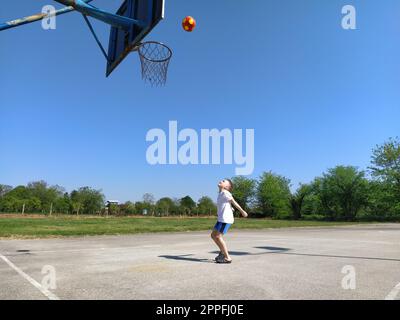 Le garçon lance la balle dans le filet de basket-ball. L'enfant joue avec une balle sur l'aire de jeux. Espace libre pour le texte. Ciel bleu en arrière-plan. Enfant aux cheveux blonds. Banque D'Images