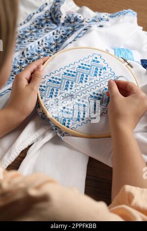 Femme brodant une chemise blanche avec fil bleu sur une table en bois, gros plan. Vêtements nationaux ukrainiens Banque D'Images