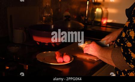 Préparer des légumes sur la cuisine. Les mains de femme senior coupant la carotte bouillie sur la plaque de plastick dans les lumières de coucher de soleil. Vieille femme cuisinant la salade Banque D'Images