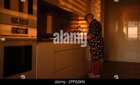 Préparer des légumes sur la cuisine moderne. Femme senior coupant la carotte bouillie sur la plaque de plastick dans les lumières du coucher du soleil. Vieille femme cuisinant la salade Banque D'Images