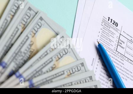 le formulaire d'impôt de 1120 se trouve près de billets de cent dollars et un stylo bleu sur un fond bleu clair. Déclaration de revenus des sociétés AMÉRICAINES Banque D'Images