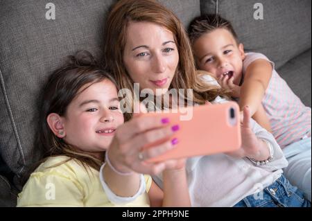 Mère heureuse avec ses enfants font un selfie ou un appel vidéo au père ou à des parents dans un canapé. Concept de technologie, nouvelle génération, famille, connexion, parentalité. Banque D'Images