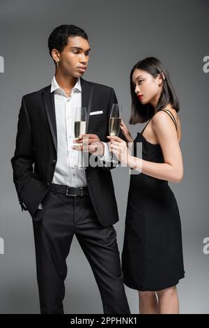 élégant africain américain homme en costume et asiatique femme en robe noire posant avec des verres de champagne isolés sur gris, image de stock Banque D'Images