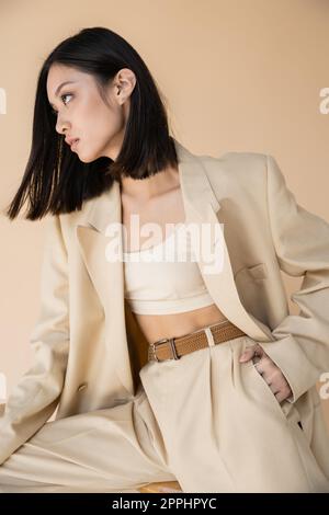 brunette asiatique femme en costume élégant posant avec la main dans la poche et regardant loin sur fond beige, image de stock Banque D'Images