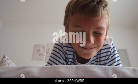 Mignon petit garçon caucasien de 10 ans se trouve à la maison sur le lit dans la chambre et sourit slyly, plissant et fermant les yeux. Attendre une surprise, une blague et se faire dorloter. Peekaboo. Jouez à Blinders. Banque D'Images