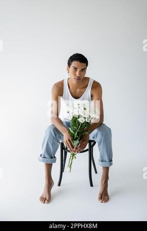 pleine longueur d'homme afro-américain en débardeur et jeans tenant des fleurs et regardant l'appareil photo tout en étant assis sur fond gris, image de stock Banque D'Images