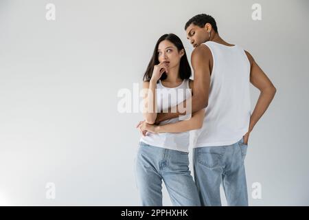 homme afro-américain en haut blanc tank et jeans embrassant la femme asiatique rêveuse regardant loin isolé sur gris, image de stock Banque D'Images