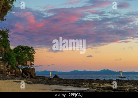 Vue sur la côte de l'île de Nosy Komba bordée de palmiers et de bateaux flottant dans la mer au coucher du soleil, Nosy Komba, Madagascar Banque D'Images