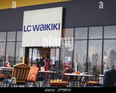 Sremska Mitrovica, Serbie, 27 février 2021 Centre commercial, magasin de vêtements LC WAIKIKI. Un homme portant un masque de protection chirurgical sort des portes coulissantes d'un magasin. Café avec chaises et tables en métal Banque D'Images