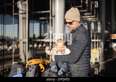 Fatherat réconfortant son bébé bébé garçon en pleurs enfant fatigué assis sur le dessus du chariot à bagages en face de la gare de l'aéroport tout en voyageant avec la famille. Banque D'Images