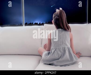 Jolie petite fille assise sur un canapé près de la fenêtre et regardant l'étoile de tir dans un beau ciel nocturne Banque D'Images