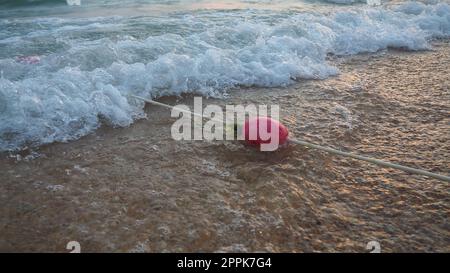 Bouées sur une corde près de l'eau de mer. Les bouées sont des attaches roses pour alerter les gens de la profondeur de l'eau. Sauvetage de la noyade. Délimitant un lieu sur une plage de sable entre les hôtels. Vague avec des bulles. Banque D'Images