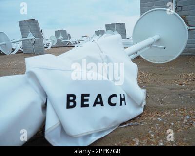 Un parasol était plié sur une plage de sable fin après le mauvais temps. Tempête en mer. Fin de la saison des plages en raison du typhon et du cyclone. Chaises longues, parasols et mobilier d'hôtel. Mot de plage écrit sur blanc Banque D'Images