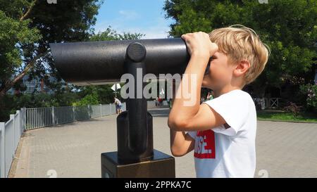 Anapa, Russie, le 23 août 2021 Un garçon regarde à travers un télescope extérieur. Un enfant caucasien de 9 ans tient un grand télescope marin avec ses mains. La rue de la station balnéaire et les touristes à pied Banque D'Images