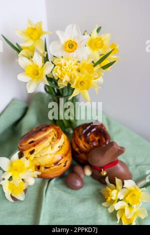 Pâtisseries traditionnelles de Pâques maison reposent sur une serviette verte avec des fleurs de jonquille, lapin, œufs en chocolat. Pâtisserie et décor de Pâques, photo verticale. Banque D'Images