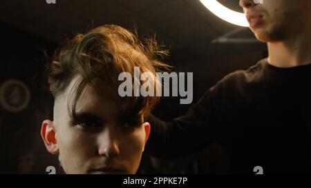 Salon de coiffure. Homme dans la chaise de barbier, coiffeur secouant les cheveux par la main avant de les couper et de les coiffer. Concept Barbershop. vue de face en éclairage sombre clé avec lumière chaude Banque D'Images