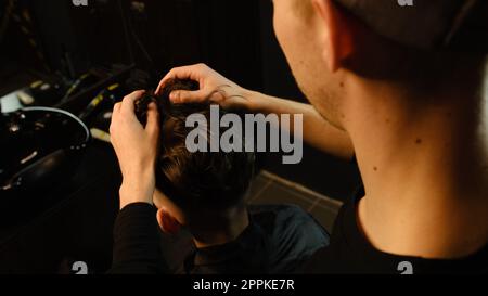 Salon de coiffure. Homme dans la chaise de barbier, coiffeur coiffant ses cheveux avec les mains dans l'éclair sombre clé. Coiffeur pour homme Banque D'Images