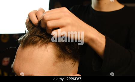 Salon de coiffure. Homme dans la chaise de barbier, coiffeur coiffant ses cheveux avec les mains dans l'éclair sombre clé. Coiffeur pour homme Banque D'Images