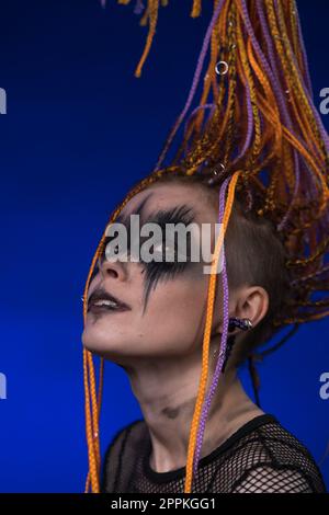 Portrait cinématographique d'une jeune femme avec maquillage noir d'horreur peint sur le visage et coiffure de Dreadlocks de couleur orange. Studio tourné sur fond bleu Banque D'Images