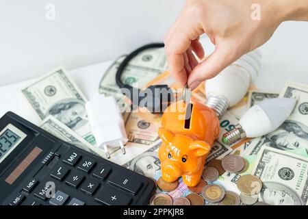 La main d'une femme met une pièce dans une tirelire orange sous la forme d'un cochon, économisant de l'argent. Calculatrice, billets de banque en euros et en dollars, pièces de monnaie. Temps d'imposition, paiement de facture, calculateur pour compter. Banque D'Images