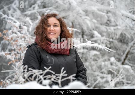 La jeune femme aux cheveux bruns est souriante et regarde la caméra tout en se tenant au milieu d'une forêt enneigée pendant l'hiver Banque D'Images