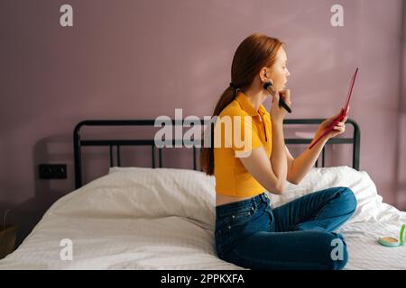 Vue latérale de la jeune femme à tête rouge concentrée regardant dans le miroir faire des soins de la peau appliquer une brosse de maquillage sur le visage et de soins de beauté assis sur le lit Banque D'Images