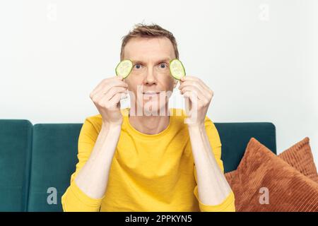 Homme adulte heureux assis sur la prise du canapé dans les mains taches oculaires de tranches de concombre frais. Masque hydratant pour gonfler. Banque D'Images