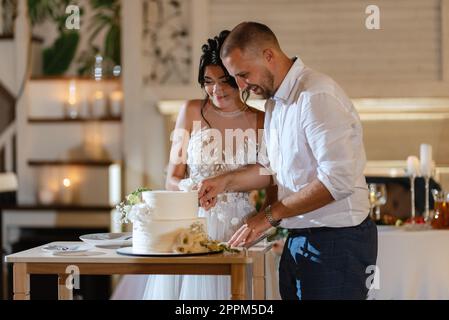 les jeunes mariés coupent joyeusement et goûtent le gâteau de mariage Banque D'Images