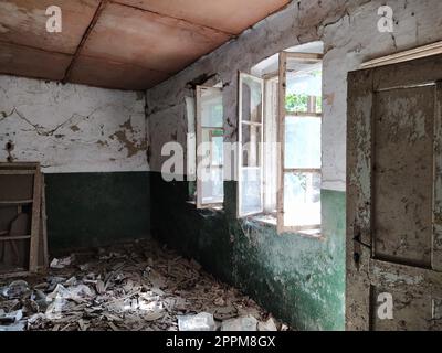 A l'intérieur d'une maison abandonnée. Des déchets éparpillés sur le sol. Peinture verte, fissures et toiles d'araignée sur le mur. La moitié supérieure blanche du mur. Ouvrez les fenêtres à travers lesquelles la lumière du soleil brille. Vieille porte Banque D'Images