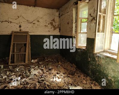 A l'intérieur de la maison abandonnée. Des déchets éparpillés sur le sol. Peinture verte, fissures et toiles d'araignée sur le mur. La moitié supérieure blanche du mur. Fenêtres ouvertes à travers lesquelles la lumière du soleil brille à travers les cadres retirés Banque D'Images