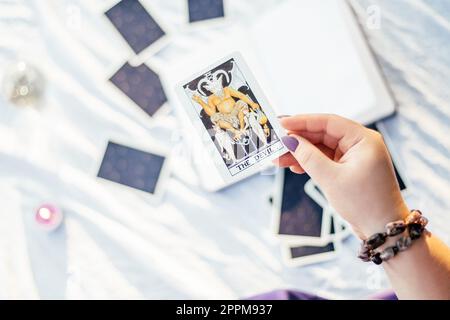 Main féminine avec des ongles violets tient la carte de tarot nommée Devil sur la surface blanche avec cahier ouvert et bougie. Vue de dessus Banque D'Images