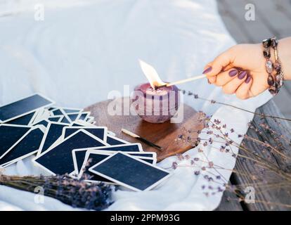 La main de la femme dans le bracelet avec de longs ongles violets allume la bougie avec allumette, à côté du jeu de cartes et de fleurs de lavande Banque D'Images