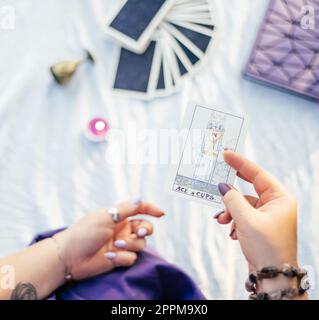 Main féminine avec des ongles violets tient la carte de tarot nommée Ace of CUPS sur la surface blanche avec cahier et bougie. Vue de dessus. Banque D'Images