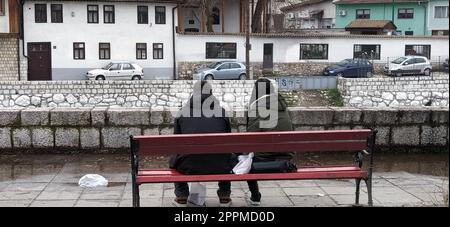 Sarajevo, Bosnie-Herzégovine - 08 mars 2020. Un jeune couple assis sur un banc en bois sous un arbre. Le remblai de la rivière Milyack. Vue sur la ville de Sarajevo, une mosquée et un minaret. Banque D'Images