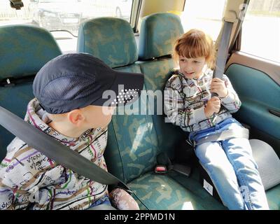 Enfants dans la voiture. Un garçon et une fille sont assis sur la banquette arrière, riant et grimacant. Les enfants attachent leur ceinture de sécurité. Un enfant dans une casquette. Jumeaux de visages différents vêtus des mêmes vêtements Banque D'Images