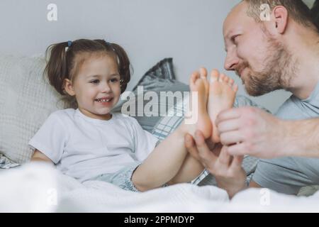 Père joue avec la petite fille 2-4 sur le lit. Papa chatouille les pieds des enfants. Famille, s'amuser Banque D'Images