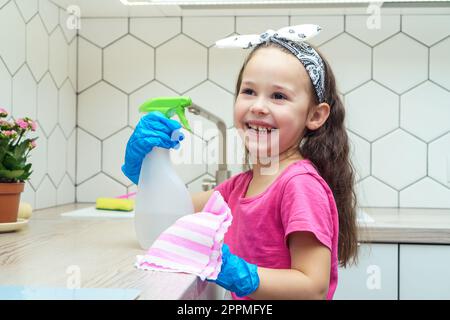 Joyeuse petite fille dans des gants de protection essuyez la cuisine avec un chiffon ménager et un pulvérisateur de détergent. Nettoyage humide à la maison. Banque D'Images