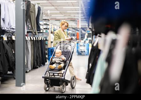 Casualy habillé mère choisissant des chaussures sportives et des produits de vêtements dans le département de sports du magasin de supermarché avec son bébé garçon enfant dans la poussette. Banque D'Images