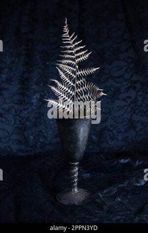 Encore la vie d'une fougères sèches frond dans une ancienne tasse d'argent terni antique contre un fond noir. Banque D'Images
