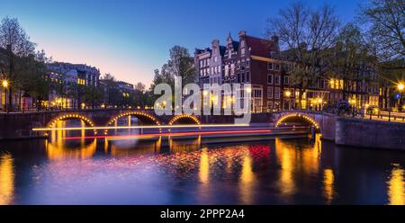 Un paysage urbain d'Amsterdam aux pays-Bas au crépuscule, éclairé par des sentiers lumineux qui réfléchit au bord de la rivière : les bateaux passent sous un pont et des bâtiments l Banque D'Images