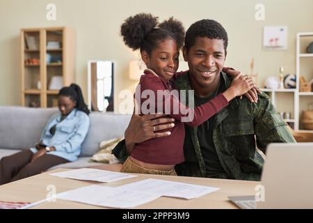 Petite fille qui embrasse son père lorsqu'il fait du travail en ligne sur un ordinateur portable à la table à la maison Banque D'Images