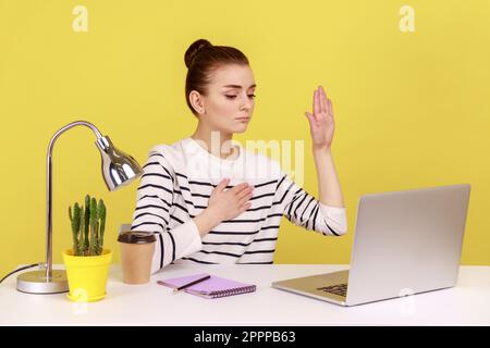 Femme honnête assise sur le lieu de travail et levant la main pour donner la promesse, regardant le lieu de travail, prêtant avec une expression fidèle digne de confiance. Studio d'intérieur tourné isolé sur fond jaune. Banque D'Images