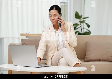Jeune femme d'affaires confiante avec un smartphone à l'oreille, assise sur un canapé devant un ordinateur portable et regardant des informations en ligne à l'écran Banque D'Images