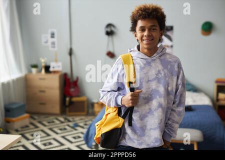 Joyeux adolescent dans un élégant sweat à capuche tenant un sac à dos jaune sur l'épaule tout en se tenant devant l'appareil photo dans sa chambre et en vous regardant Banque D'Images