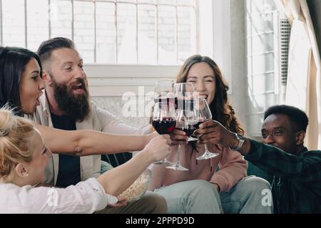 Santé ! Les gens célèbrent et élèvent des verres de vin rouge pour des toasts. Concentrez-vous sur les lunettes. Groupe d'hommes et de femmes heureux qui applaudissent et s'amusent à l'intérieur de liv Banque D'Images