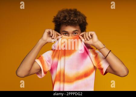 Un adolescent mignon se cache derrière un t-shirt coloré tout en se tenant devant l'appareil photo sur fond jaune pendant la séance photo Banque D'Images