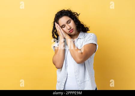 Portrait d'une femme fatiguée et exténuée avec des cheveux ondulés et foncés qui dorment sur les paumes, qui a une sieste confortable et se repose. Studio d'intérieur isolé sur fond jaune. Banque D'Images