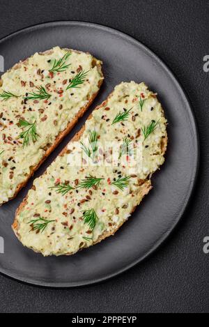 Toast grillé avec pâte d'avocat, œufs, fromage à la crème, sel, épices et herbes sur une table en béton texturé Banque D'Images