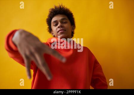 Jeune Afro-américain gars à capuche rouge regardant l'appareil photo tout en se tenant debout sur fond jaune et en gardant la main droite devant lui Banque D'Images